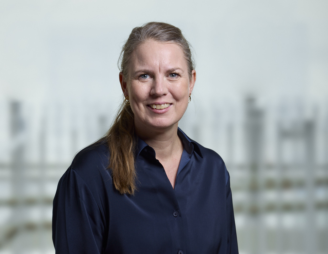 Christina E. Wanscher i et professionelt portræt med mørkeblå skjorte og lys, sløret baggrund.