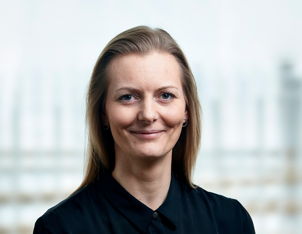 Smilende kvinde med lyst hår i mørk skjorte på en lys, sløret baggrund.