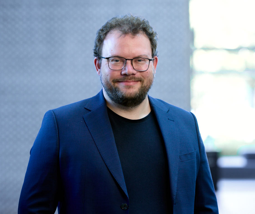 Smilende mand med briller, skæg, blå blazer og mørk trøje i et professionelt portræt indendørs.