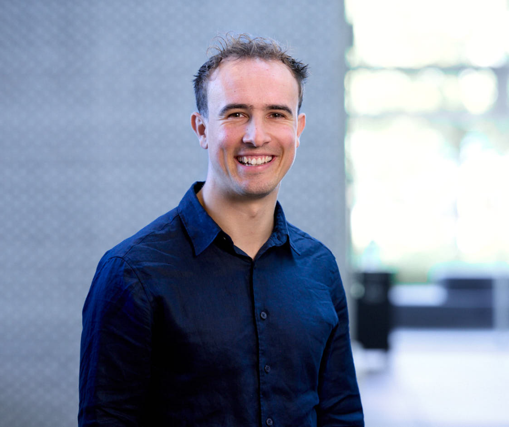 Smiling man in a dark blue button-up shirt standing indoors.