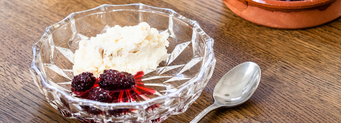 Glasskål med hvid dessert og mørke bær i rød sauce på et træbord med en ske ved siden af.