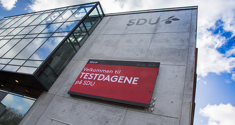 Lavvinklet billede af en SDU-bygning med glasfacade og et rødt skilt med teksten "Velkommen til TESTDAGENE på SDU".