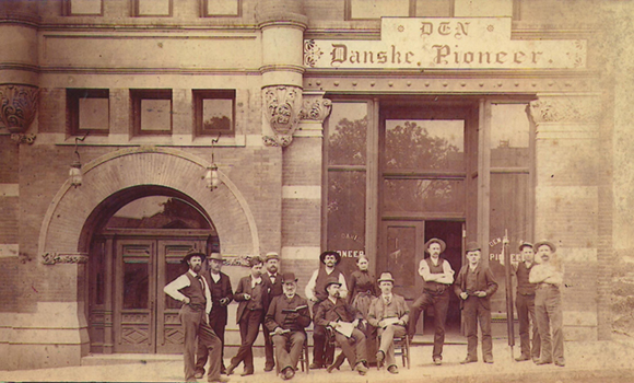 Gammelt sepia-billede af bygningen, der huser Den Danske Pioneer, som var en dansk avis i Omaha, Nebraska. Foran bygningen står en række mænd. 