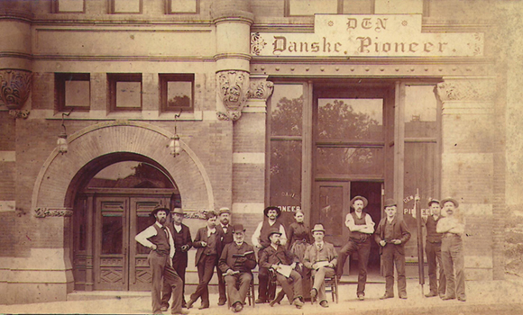 Gammelt sepia-billede af bygningen, der huser Den Danske Pioneer, som var en dansk avis i Omaha, Nebraska. Foran bygningen står en række mænd.