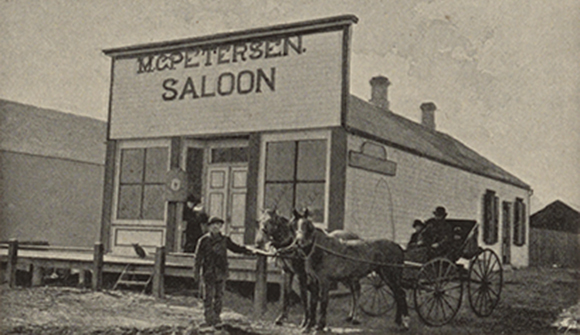 Sort-hvid billede af M.C. Petersens saloon i den lille landsby Dannebrog i Nebraska. Foran saloonen står en mand og holder to heste, der er spændt fast til en hestevogn. 