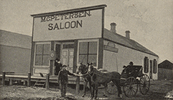 Sort-hvid billede af M.C. Petersens saloon i den lille landsby Dannebrog i Nebraska. Foran saloonen står en mand og holder to heste, der er spændt fast til en hestevogn.