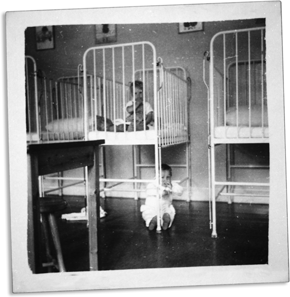 Sort-hvid billede af en termmeseng og to børn. Det ene barn befinder sig sengen, mens det andet barn sidder på gulvet og holder om sengens ben. 