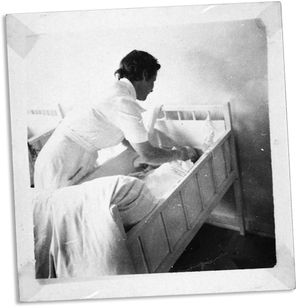 Sort-hvid billede af en plejerske, der rækker ned i en seng efter et barn.