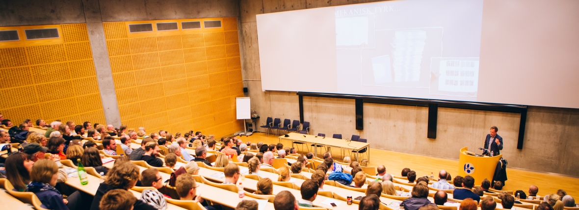 Fyldt auditorium med publikum og en oplægsholder foran et stort lærred.