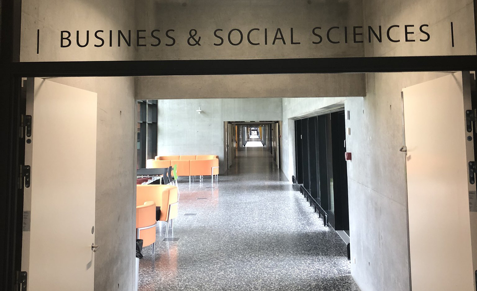Åben indgang til en moderne gang med skiltet BUSINESS & SOCIAL SCIENCES over døren og orange møbler langs korridoren.