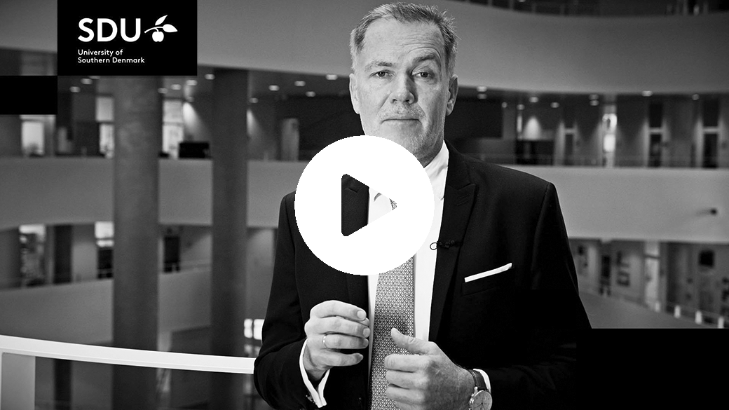 Mand i jakkesæt i et indendørs atrium med "SDU" og "University of Southern Denmark" øverst til venstre samt et stort afspilningsikon i midten.