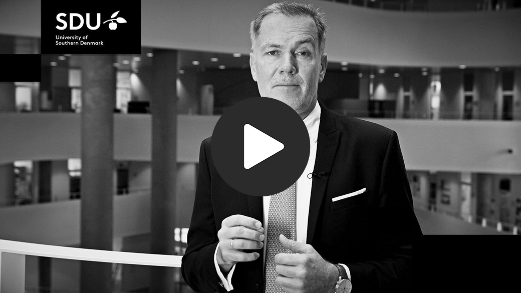 Sort-hvid videominiature med en mand i jakkesæt i et atrium, SDU-logoet med teksten "University of Southern Denmark" øverst til venstre og et stort afspilningsikon i midten.