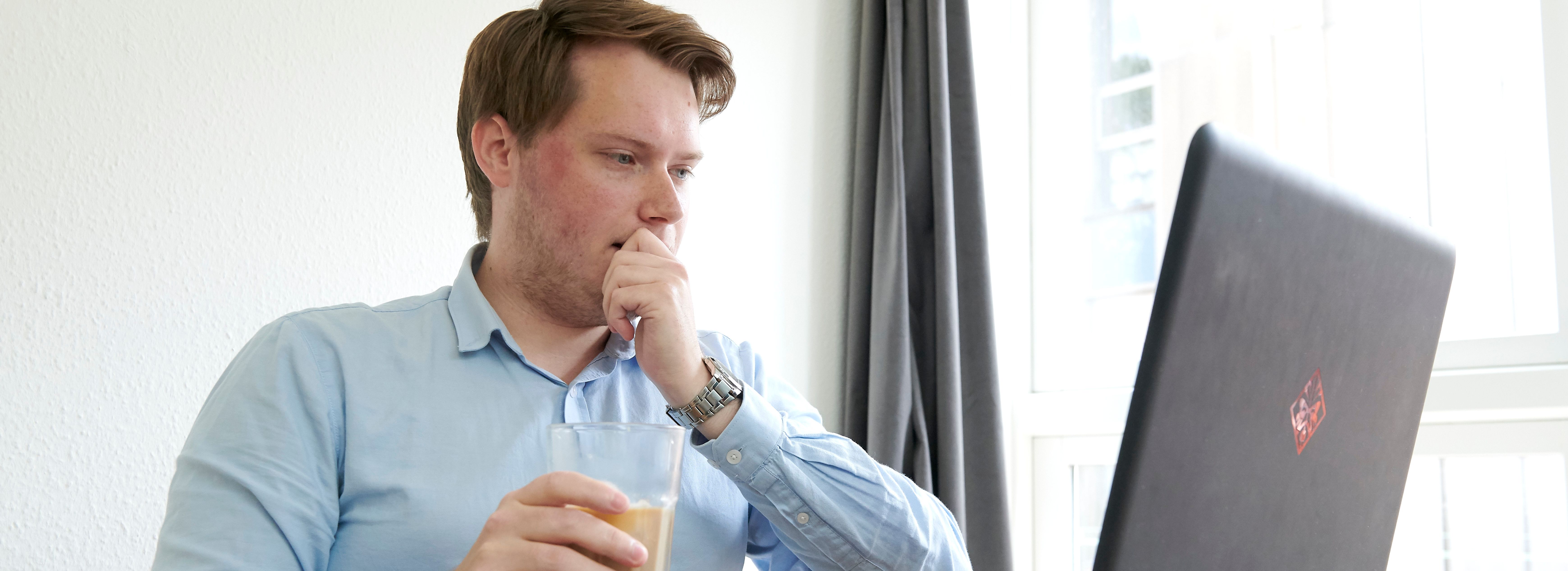 Ung mand i lyseblå skjorte ser koncentreret på en bærbar computer og holder et glas.