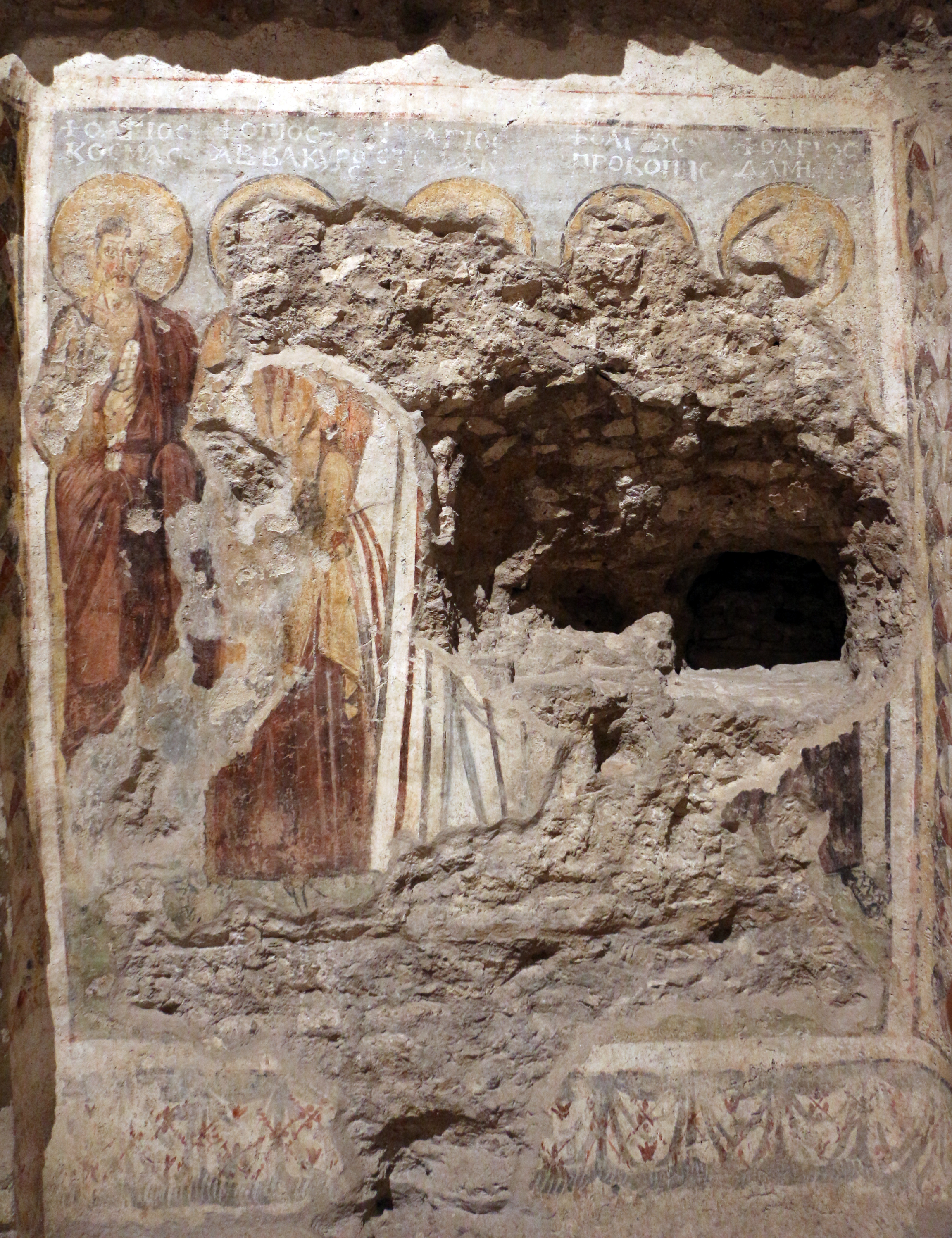 Ancient damaged fresco with haloed figures, missing plaster, a central cavity, and faded Greek text along the top.