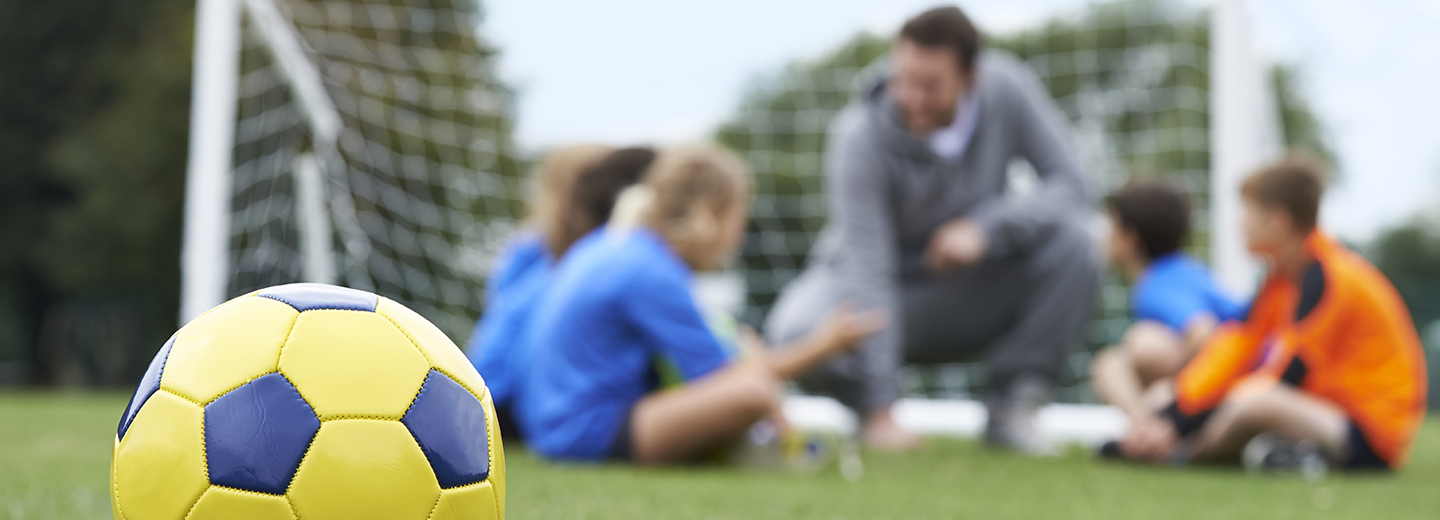 Gul og blå fodbold på græs foran en træner og børn ved et fodboldmål.