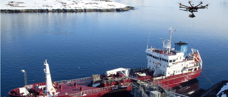 Multi-rotor drone flying over a red ship docked in calm coastal water beside a snowy shoreline.