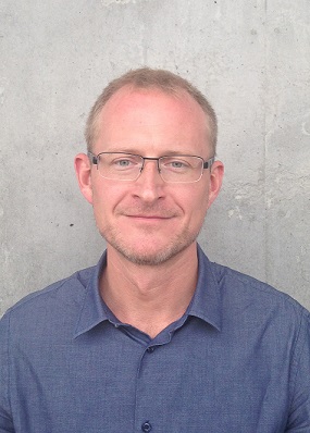 Man wearing glasses and a blue shirt in a portrait against a concrete wall.