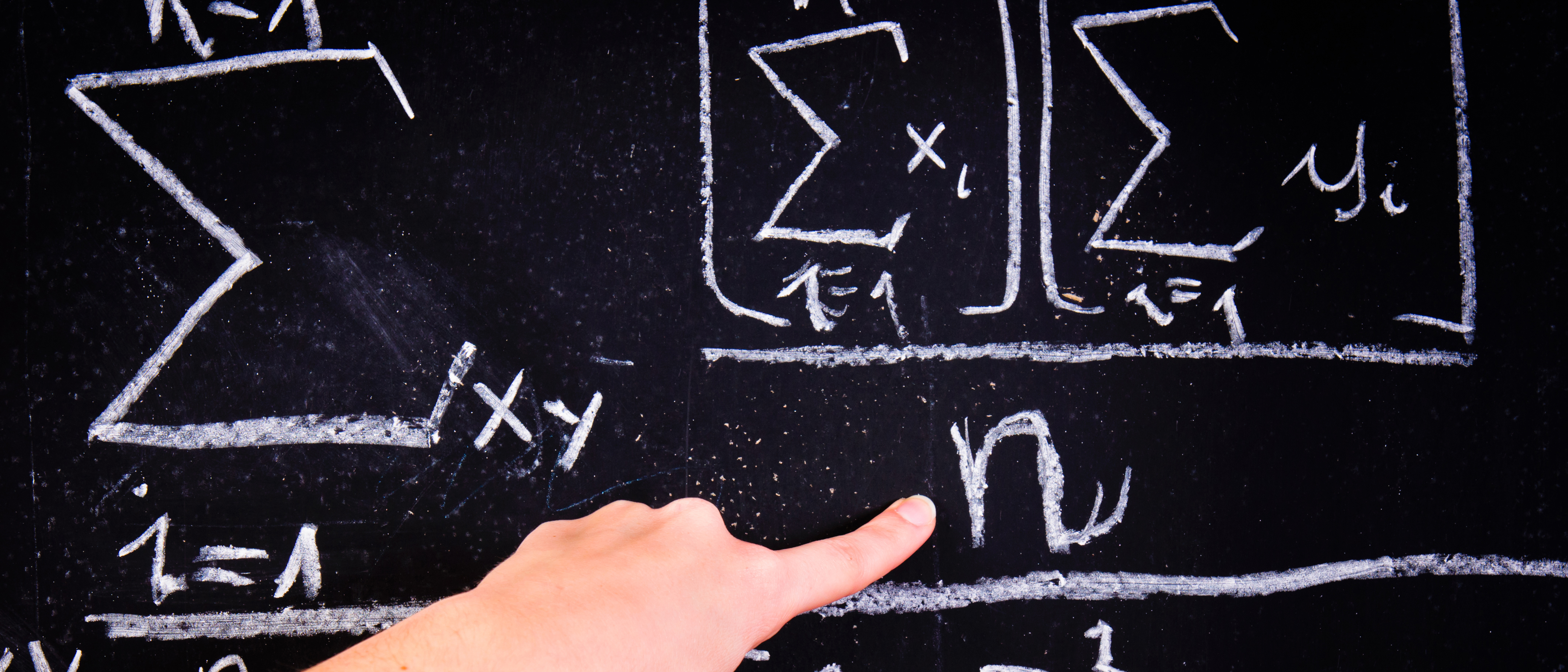Close-up of a hand pointing at chalk-written mathematical symbols on a blackboard, with visible text "xy", "1=1", and "yi".