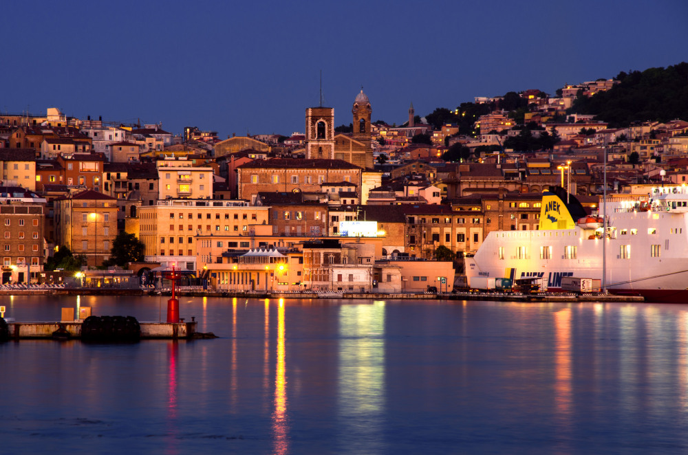 Twilight waterfront cityscape with hillside buildings, church towers, reflections on calm water, and a ferry displaying "ANEK" and "LINES".