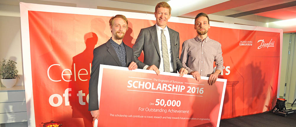 Tre mænd med et stort skilt foran en rød Danfoss-baggrund med teksten “SCHOLARSHIP 2016”, “DKK 50,000” og “For Outstanding Achievement”.