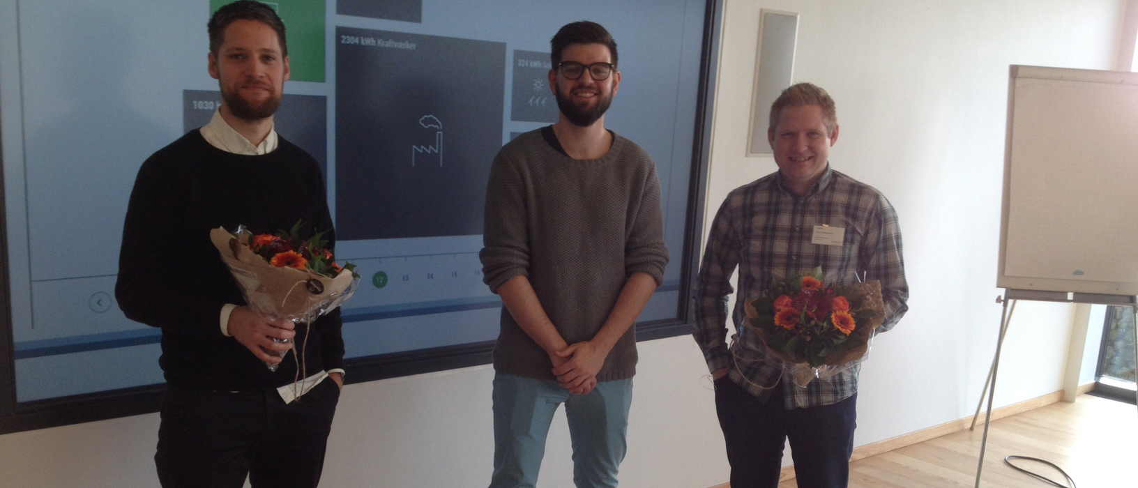 Three men posing indoors in front of a large display screen, with two holding bouquets of flowers and presentation text including "2396 kWh fabrikker" visible in the background.