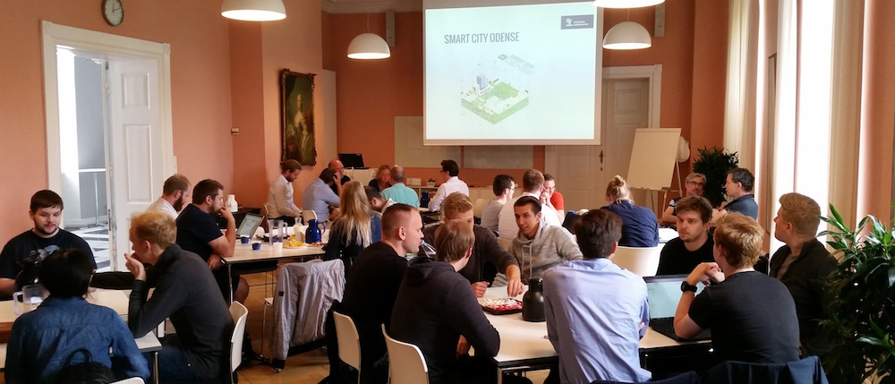 Deltagere ved borde i et mødelokale under en præsentation med teksten "SMART CITY ODENSE" på et stort lærred.