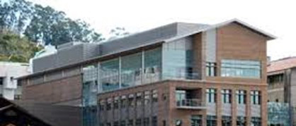Modern multi-story brick and glass building exterior with large windows and trees behind it.