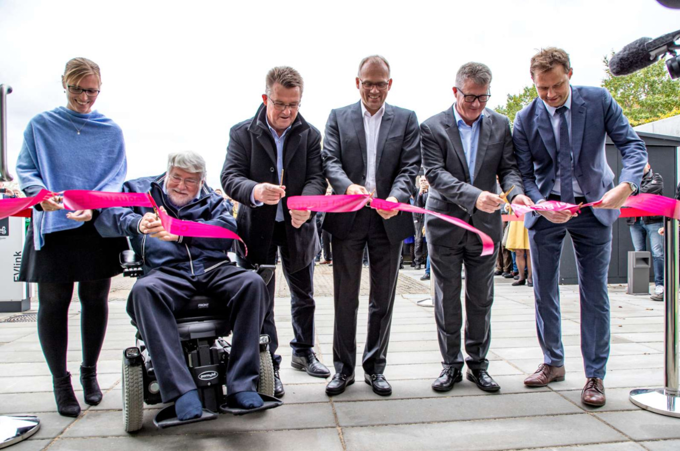 Six participants cut a bright pink ribbon at an outdoor inauguration, with one person seated in a powered wheelchair.