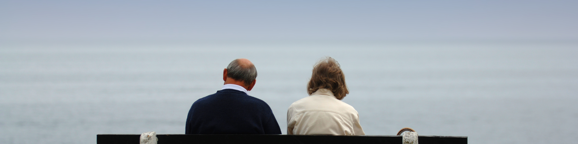 Two older adults seen from behind sitting on a bench by the sea.