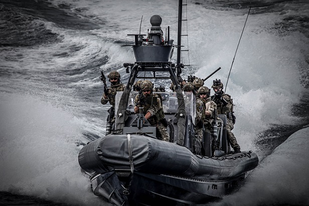 Armed military personnel on a rigid inflatable boat speeding through rough water.