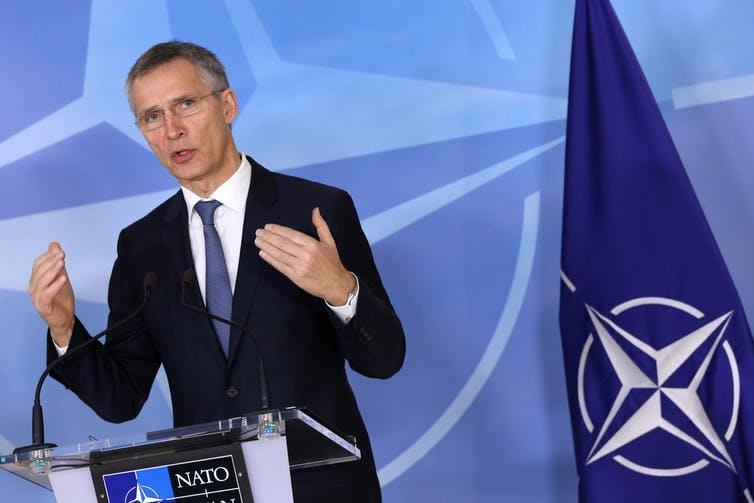 Man speaking at a podium labeled "NATO" beside a NATO flag and blue backdrop.