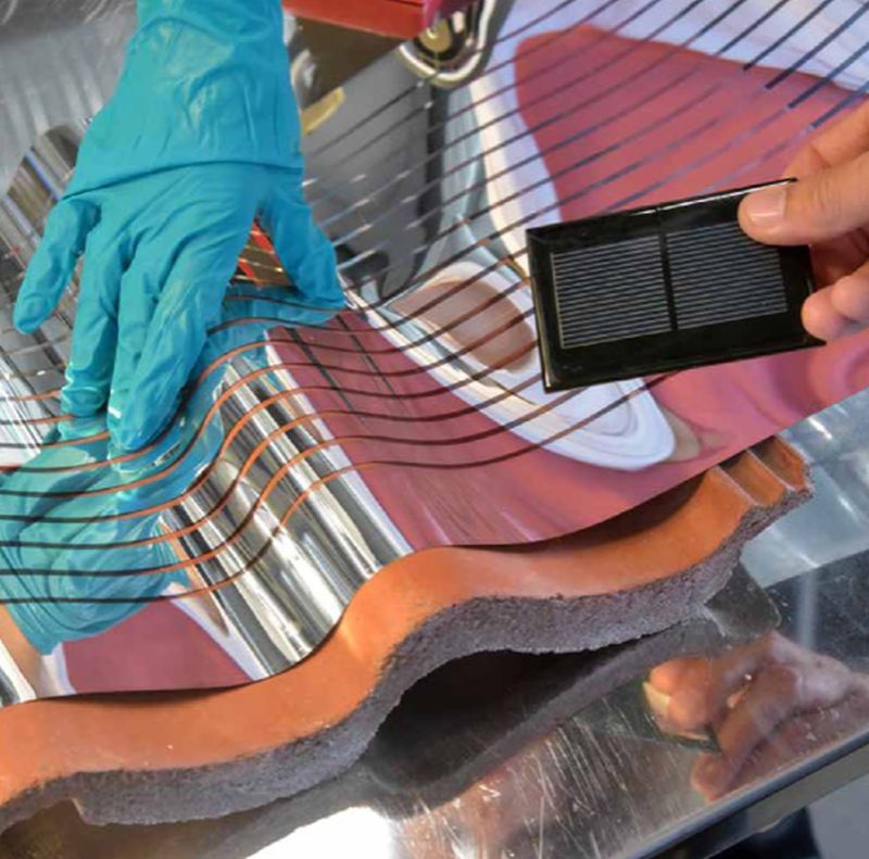 Blå handske og hånd placerer fleksibelt solcellemateriale og en lille sort solcelle på en bølget tagplade.