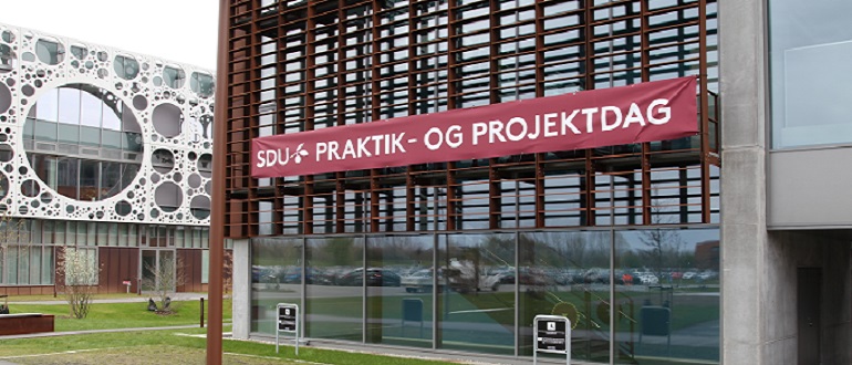 Moderne bygning med et rødt banner med teksten "SDU+ PRAKTIK- OG PROJEKTDAG" på facaden.