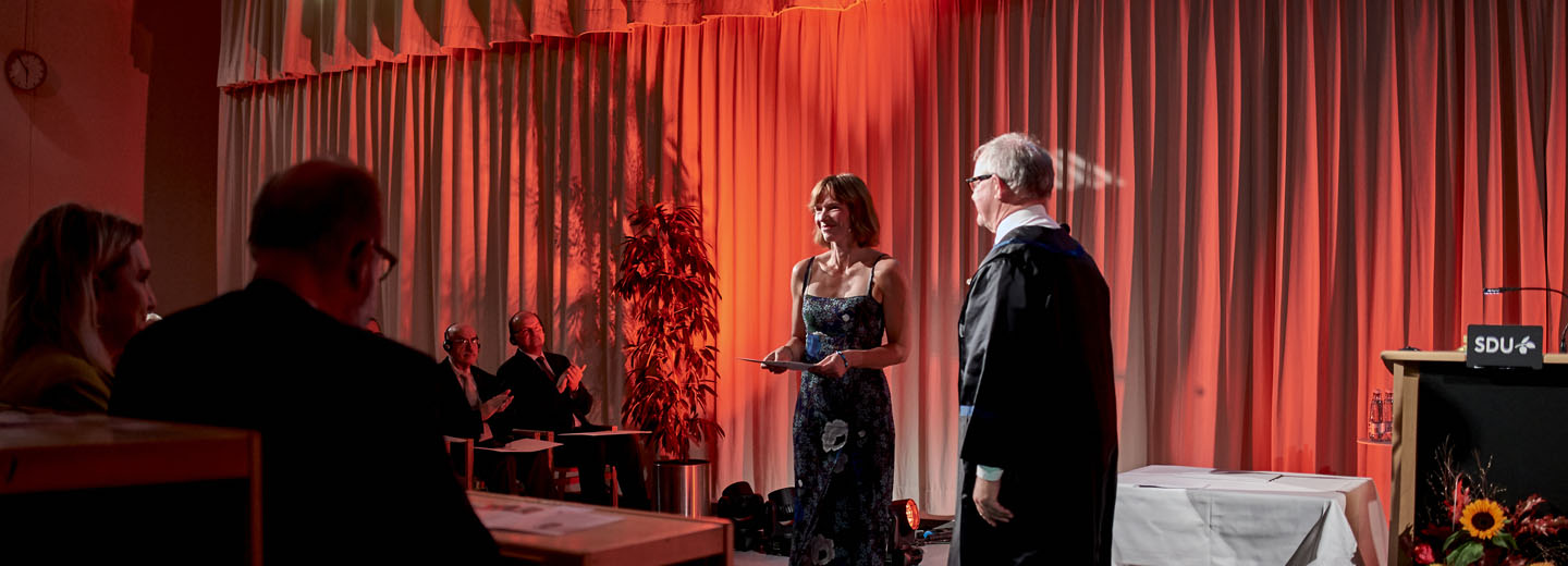 Kvinde på scene ved ceremoni med en person i akademisk kappe og talerstol med teksten "SDU".