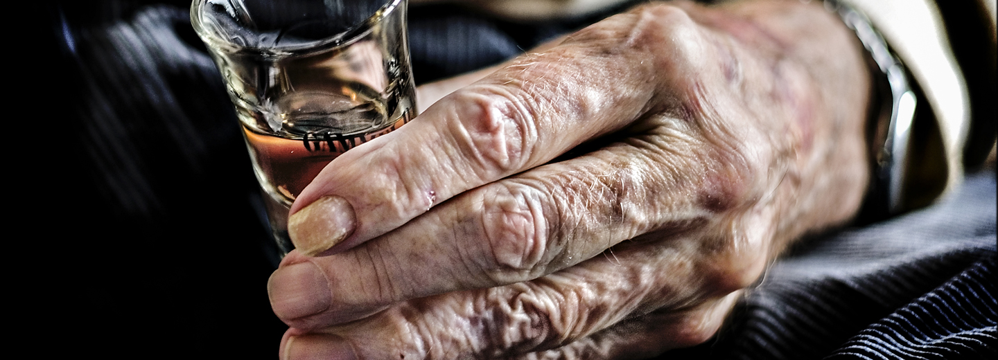 Nærbillede af en ældre hånd, der holder et lille glas med alkohol.