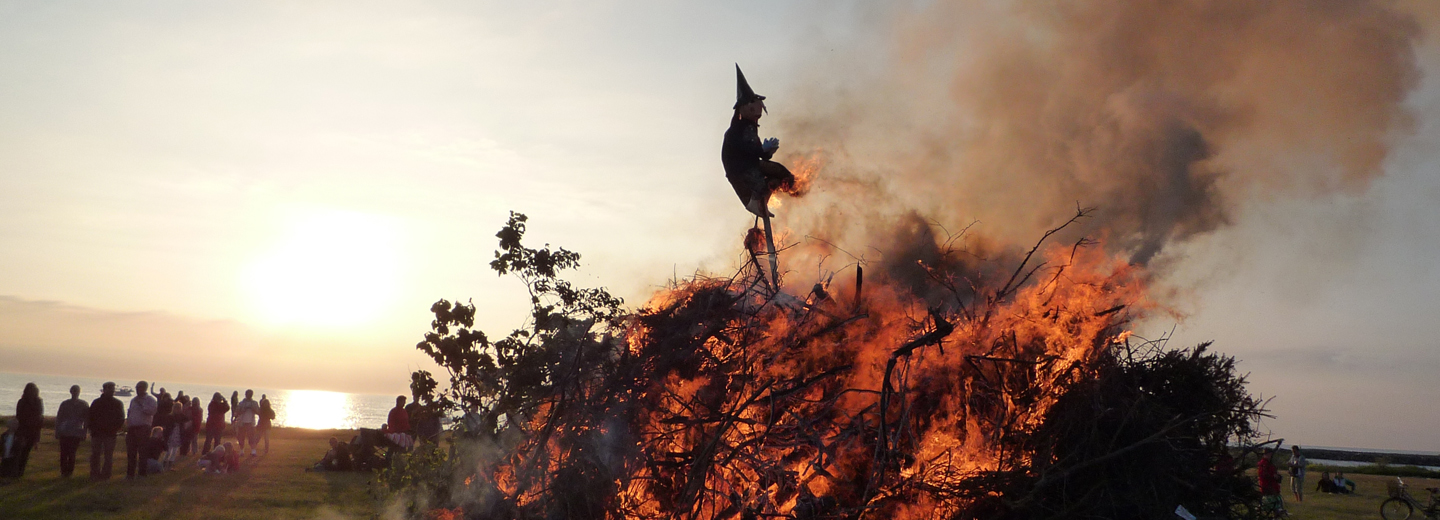 Sankt Hans-bål med heksedukke i flammer ved solnedgang og tilskuere ved kysten.