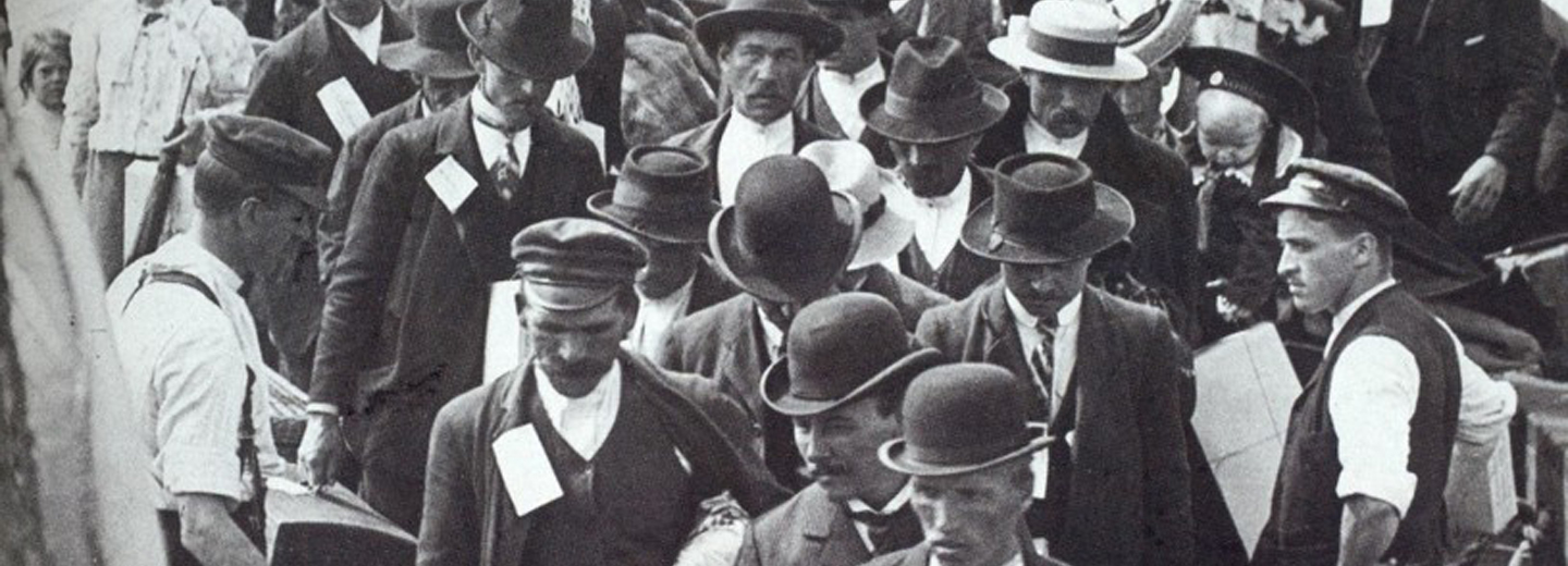 Historisk sort-hvidt foto af en tæt menneskemængde med mænd i hatte, bagage og papirer, set ovenfra.
