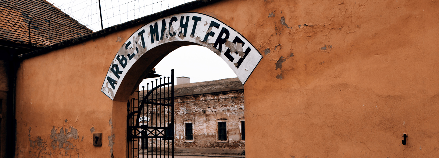 Murport med jernlåge og teksten "ARBEIT MACHT FREI" over indgangen, med en bygning og pigtråd i baggrunden.