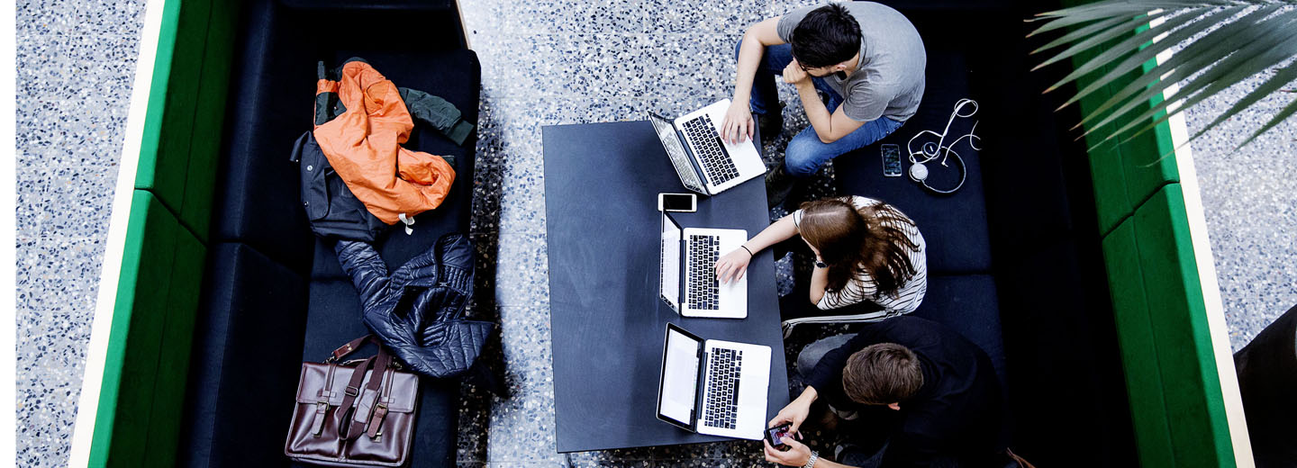 Tre personer arbejder på bærbare computere ved et bord set ovenfra i et loungeområde med jakker og taske på bænken overfor.