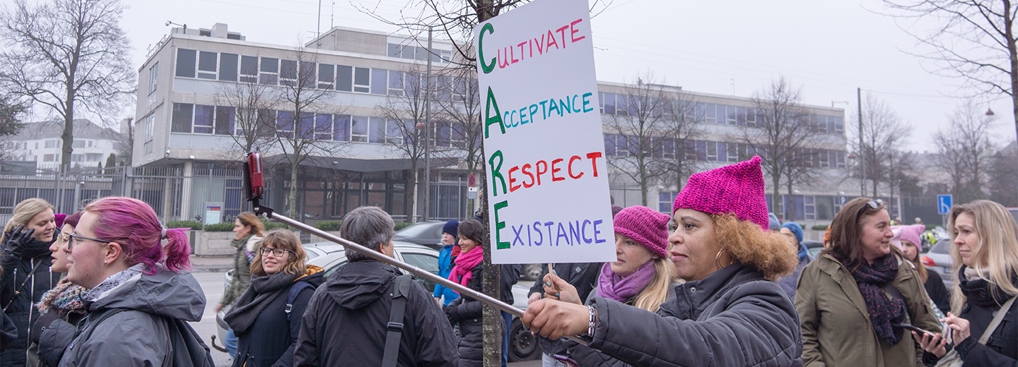Person i en lyserød hue holder et demonstrationsskilt med teksten "Cultivate Acceptance Respect Existance", omgivet af en gruppe mennesker på en gade.