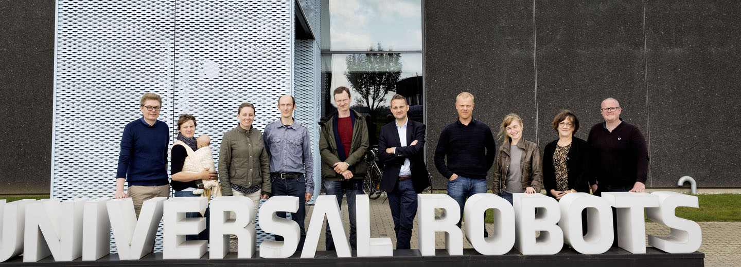 Gruppe mennesker poser foran store bogstaver med teksten UNIVERSAL ROBOTS udenfor en moderne bygning.
