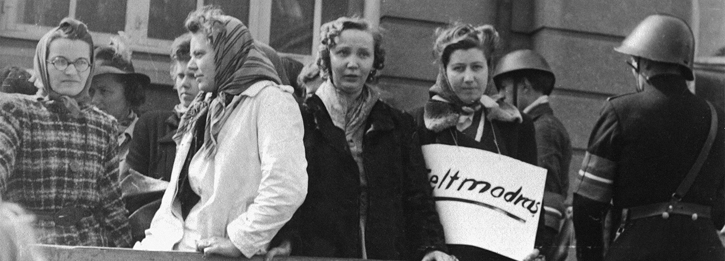 Sort-hvidt foto af en gruppe kvinder på en gade med uniformerede mænd i baggrunden og et skilt med delvist synlig tekst.