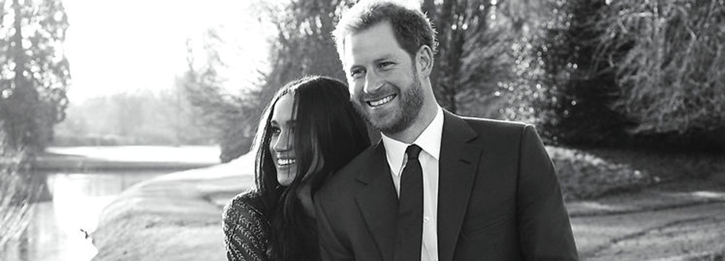 Meghan og Harry smiler sammen i et sort-hvidt udendørs portræt ved vand og træer.