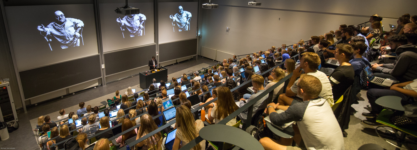 Stort auditorium med mange studerende ved computere, en underviser ved talerstolen og sort-hvide billeder projiceret på tre store skærme.