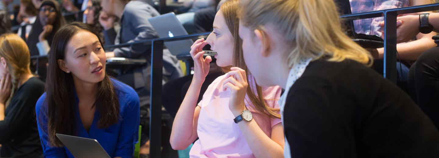 Studerende, der taler sammen i et auditorium, hvor en person holder en bærbar computer.