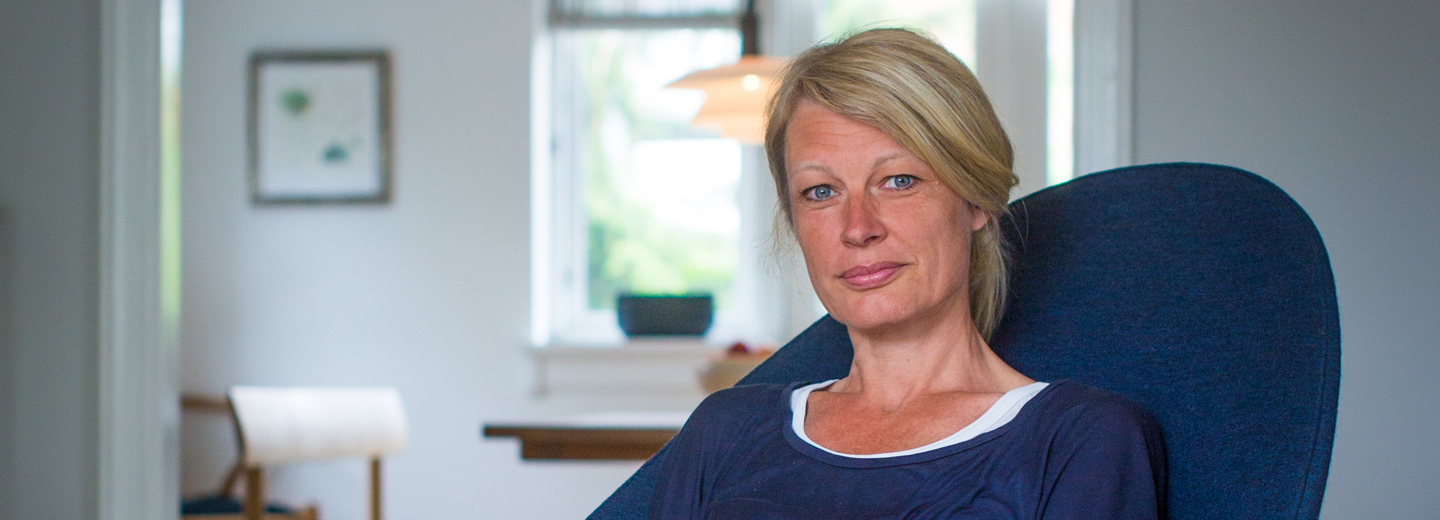 Kvinde med lyst hår sidder i en mørkeblå stol i et lyst rum.
