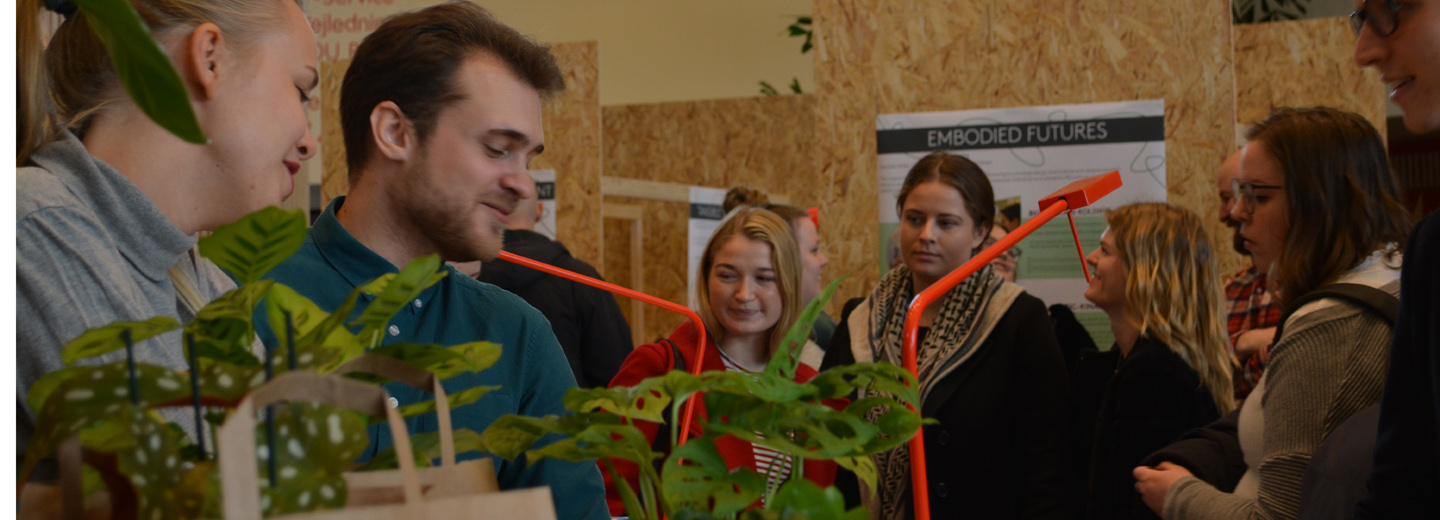 Studerende ved en udstilling indendørs med planter i forgrunden og en plakat med teksten "EMBODIED FUTURES" i baggrunden.