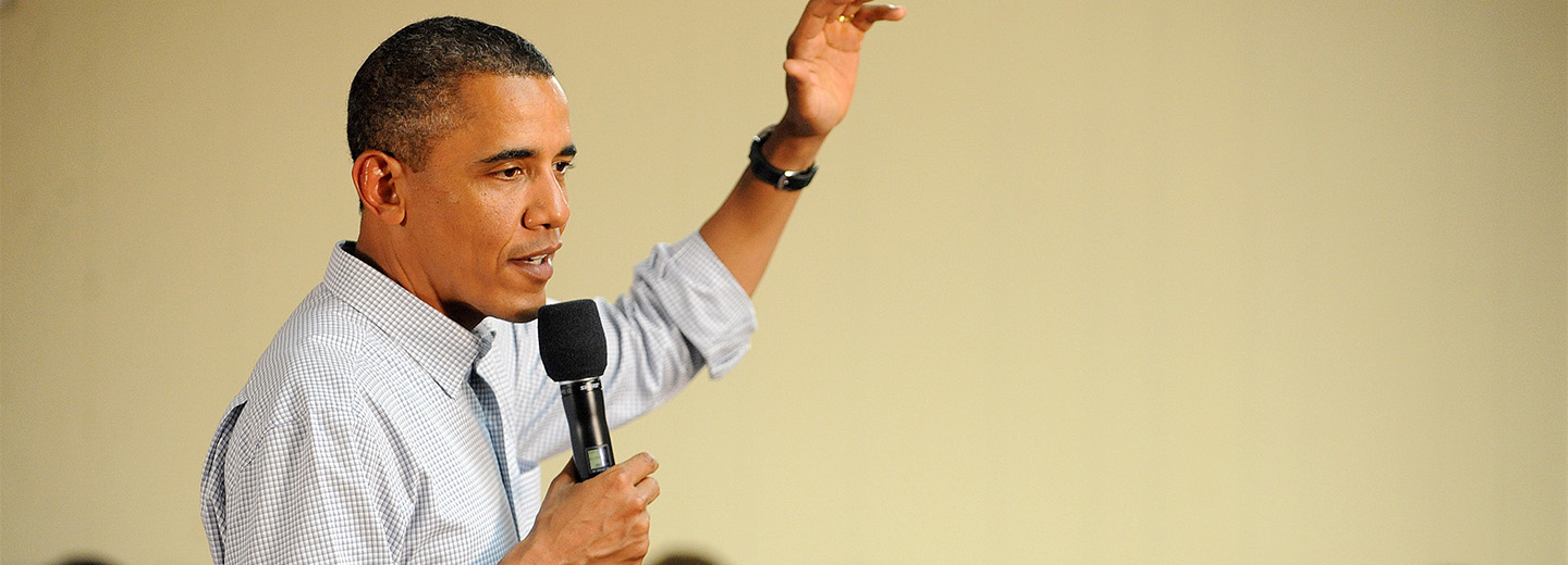 Barack Obama holder en mikrofon og taler med løftet hånd foran en lys baggrund.