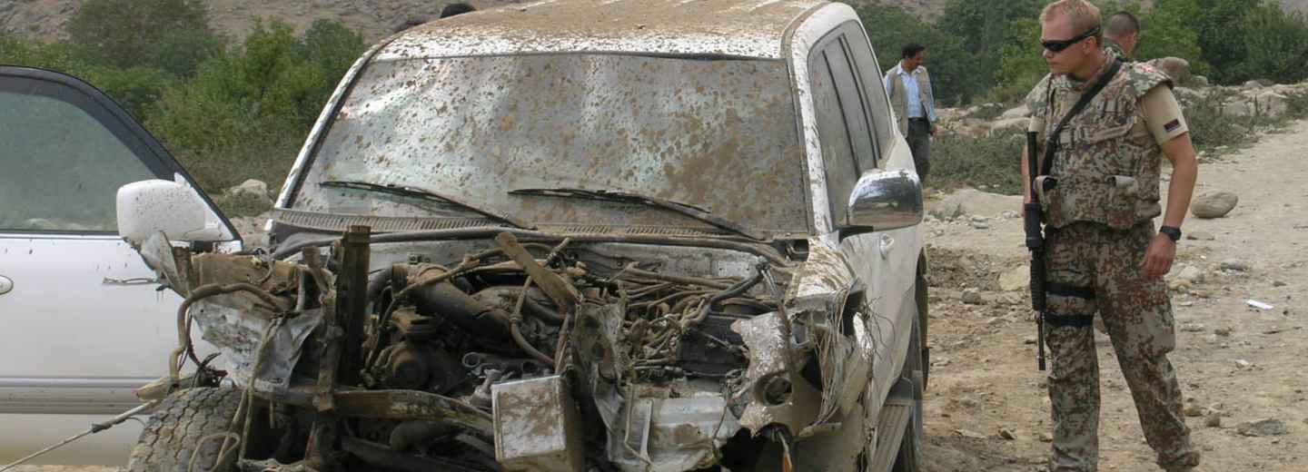 Heavily damaged white SUV on a dirt roadside with an armed soldier standing next to it.