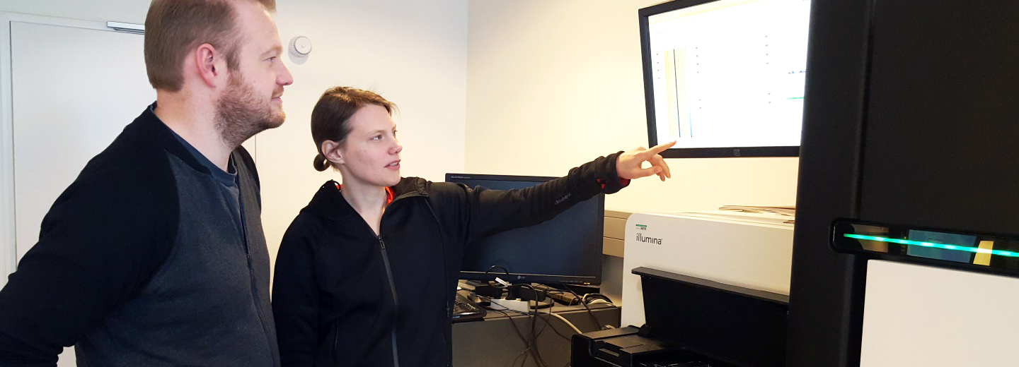 To personer i et laboratorie står ved "illumina"-udstyr, mens den ene peger på en skærm.